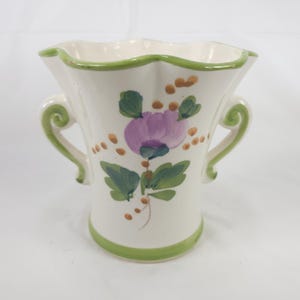 May include: A cream-colored ceramic vase with a scalloped rim and two side handles. The vase features a hand-painted design of a purple flower with green leaves and small brown dots scattered around the floral motif. The rim and base have green trim.