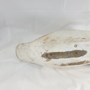 May include: A vintage, weathered duck decoy, painted white with visible wear and tear. The wooden decoy shows signs of age, with exposed wood and a distressed finish. The underside features a rectangular area with two holes.