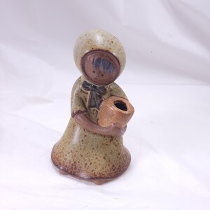 May include: A ceramic figurine of a person wearing a hooded dress and holding a small pot. The figurine is primarily tan with brown accents and a speckled finish. The pot is a lighter shade of brown.