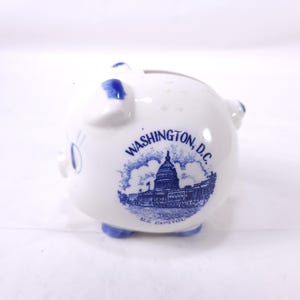 May include: White ceramic piggy bank with blue accents. The bank features a blue illustration of the U.S. Capitol building and the text "WASHINGTON, D.C." and "U.S. CAPITOL". The piggy bank has blue feet and blue accents on the ears.