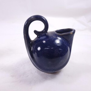 May include: A dark blue ceramic pitcher with a unique, curved handle and a spout. The pitcher has a textured surface and a glossy finish. The handle forms a complete loop, adding to its distinctive design. The overall shape is rounded, with a small opening for pouring.
