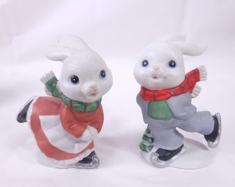 May include: Two ceramic figurines of white rabbits in winter attire. One rabbit wears a red and white dress with a green scarf, the other wears a gray suit and red scarf, both with ice skates. The figurines have blue eyes.