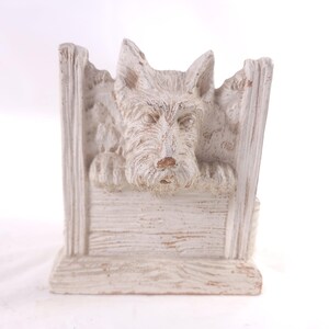 May include: A cream-colored, decorative dog sculpture. The sculpture features a Scottish Terrier dog peering over a wooden fence. The dog's fur is textured, and the sculpture has a distressed finish, giving it a rustic appearance.