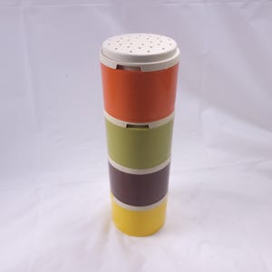 May include: A multi-colored, cylindrical container with a white, perforated lid. The container is composed of stacked sections in orange, olive green, burgundy, and yellow. The sections are separated by thin, off-white bands.