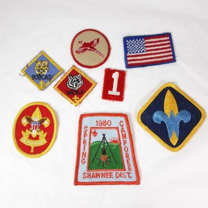May include: A collection of eight vintage Boy Scout patches, including a red, white, and blue American flag patch, a blue patch with a fleur-de-lis, a red patch with a scout emblem, a tan patch with a bird, a blue patch with a wolf, a blue patch with a bobcat, a red patch with the number 1, and a blue patch with the text "1980 Spring Camporee Shawnee Dist."