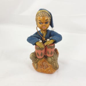 May include: A figurine of a person with a wood-grain finish wearing a blue cap and blue shirt, sitting on a brown base while playing two pink bongo drums with diamond patterns. The figure features gold trim, white pearl-like accents, and gold cord.