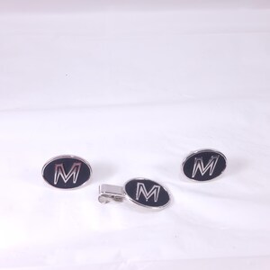 May include: A set of silver-toned metal cufflinks and a tie clip. Each piece features a black oval with a silver letter "M" in the center. The cufflinks and tie clip are displayed on a white surface.