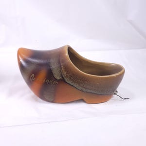 May include: A ceramic clog-shaped planter in shades of brown and orange. The planter has a glossy finish and a textured appearance. The text "O. inken" is written on the side. A small black string is attached to the back of the clog.