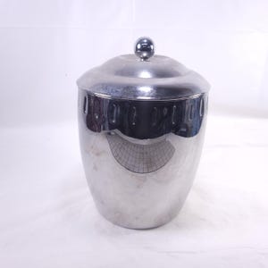 May include: A shiny, silver-colored metal container with a lid and a small, round knob on top. The container has a cylindrical shape with a slightly wider base and a series of vertical cutouts near the top. The lid is curved, and the surface reflects the surrounding environment.