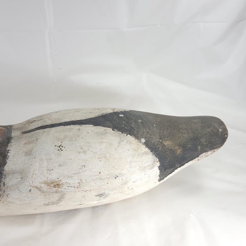 May include: A vintage duck decoy, painted in white, black, and red. The wooden decoy has a weathered appearance, with visible paint wear and a rounded shape. The head is painted black, and the body is white.