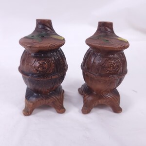 May include: A pair of brown ceramic salt and pepper shakers. Each shaker is shaped like a small stove with a rounded body, a flared top, and three small feet. The shakers have a textured surface and are painted with green and yellow accents.