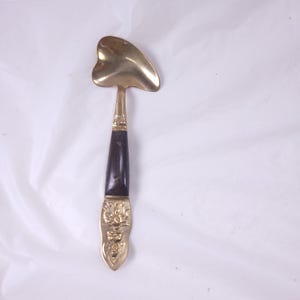 May include: A vintage brass serving spoon with a heart-shaped head and a dark wooden handle. The handle is adorned with decorative brass elements, including an ornate design at the base. The spoon is likely used for serving food.