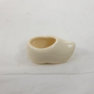 May include: A small, off-white ceramic clog, a traditional Dutch shoe. The miniature shoe has a rounded toe and a hollow interior, suggesting it could be used as a small planter or decorative item. The clog is set against a plain background.