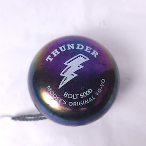 May include: A metallic purple yo-yo with the word "THUNDER" above a white lightning bolt graphic. Below the graphic, the text reads "MOOSE'S ORIGINAL YO-YO BOLT 5000". A gray string is attached.