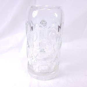 May include: Clear glass beer stein with embossed design and the words "Hofbrauhaus Munchen". The stein has a cylindrical shape with a thick base and a wide opening. The design includes a crest and floral elements.