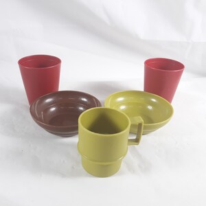 May include: A collection of vintage plastic dishware in various colors. The set includes two red cups, two bowls in brown and green, and a green mug with a handle. The items are arranged on a white surface.
