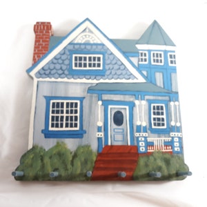 May include: A blue and white painted wooden house key holder with a red brick chimney and a red porch. The house has a white picket fence and a red door with a white oval window. There are five hooks on the bottom of the house.
