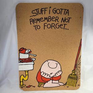 May include: A rectangular corkboard with the text "...STUFF I GOTTA REMEMBER NOT TO FORGET...". The board features a cartoon illustration of a person, a sink with dishes, and a broom. The person is wearing a red shirt.
