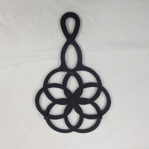 May include: Black metal trivet with an intricate, interlocking design. The trivet features a handle and a circular pattern of overlapping shapes, providing a decorative and functional element for protecting surfaces from hot cookware.