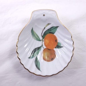 May include: White seashell-shaped dish with a gold rim. The dish features a painted design of two oranges and green leaves. The dish has a small hole at the top. A decorative piece for the home.