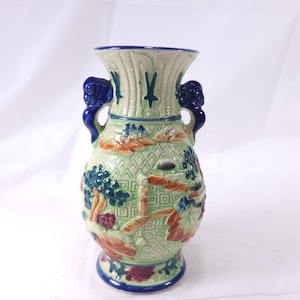 May include: A decorative ceramic vase with a light green background and blue accents. The vase features a landscape design with trees, mountains, and a bridge. It has two blue handles and a blue rim. The vase is approximately 10 inches tall.