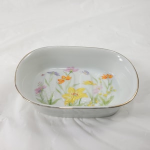 May include: An oval white ceramic soap dish featuring a delicate floral pattern with yellow, orange, and purple flowers on the interior base. The rim of the dish is finished with a thin metallic silver band, and the interior has three raised ridges.