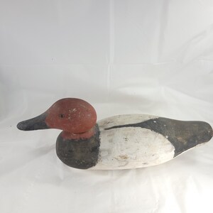 May include: A wooden duck decoy with a red head and black beak. The body is painted white with black and gray accents. The decoy is designed to float on water and attract other ducks.