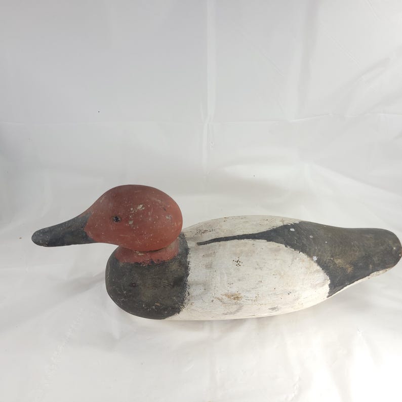 May include: A wooden duck decoy with a red head and black beak. The body is painted white with black and gray accents. The decoy is designed to float on water and attract other ducks.