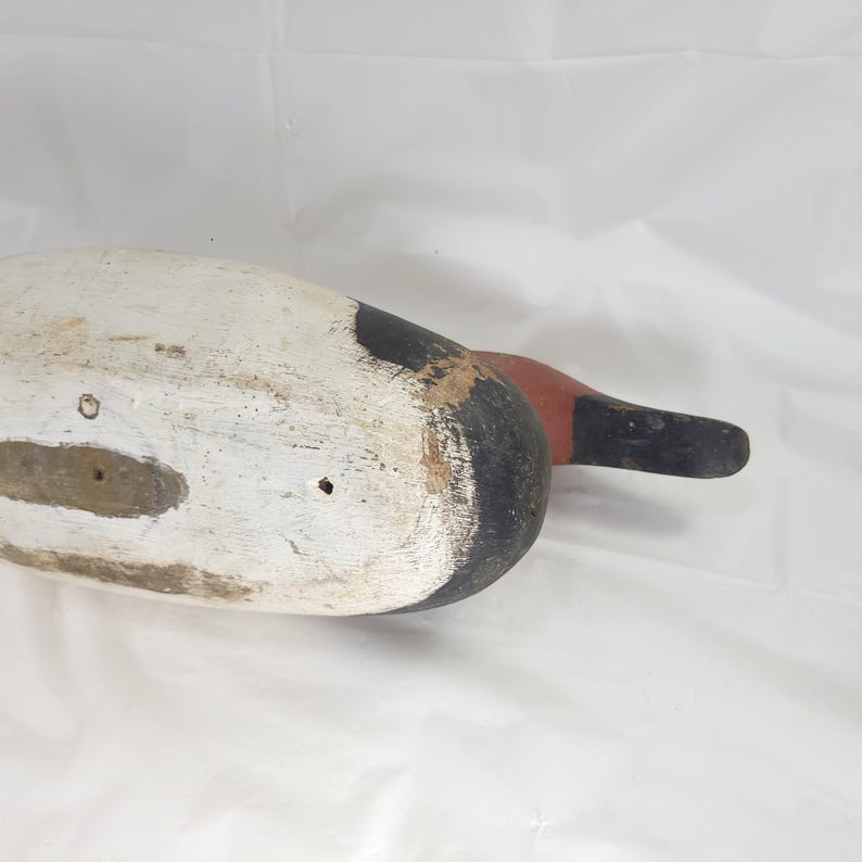 May include: A vintage wooden duck decoy, painted in white, black, and red. The decoy has a weathered appearance, with visible wear and tear. The duck's head is painted black and red, with a white body. A classic piece of folk art.