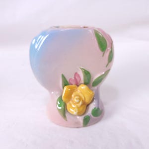 May include: A small, ceramic vase with a pastel gradient of blue, pink, and white. The vase is decorated with a yellow rose, pink buds, and green leaves. The vase has a rounded shape.