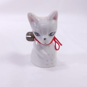 May include: A small, white ceramic cat figurine with a red ribbon tied around its neck and a small bell. The cat has detailed facial features and is sitting upright. The figurine is a decorative item.