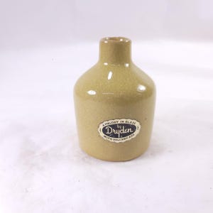 May include: A small, tan-colored ceramic bottle with a rounded body and narrow neck. The bottle has a label that reads "A Melody in Glaze by Dryden with Volcanic Ash."