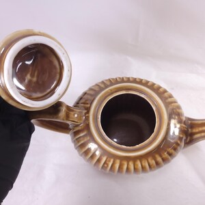 May include: A brown ceramic teapot with a matching lid. The teapot has a ribbed design around the body and a handle on the side. The lid is off, revealing the interior. The teapot is a warm brown color.