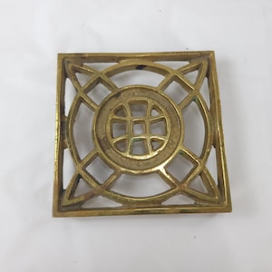 May include: A square, brass-colored trivet with an openwork design. The center features a circular pattern with a grid, surrounded by intersecting lines and curves. The trivet is designed to protect surfaces from hot items.