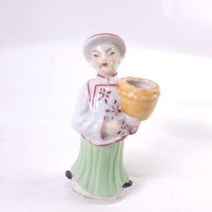 May include: A small porcelain figurine of a person in traditional Chinese attire. The figure wears a white top with red trim and floral accents, a light green skirt, and a red-trimmed hat. The figure holds a small, tan basket.