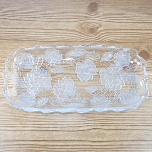 May include: A rectangular clear glass serving tray featuring a frosted floral pattern with etched flowers and leaves. The tray has a scalloped, wavy edge design and a smooth, transparent surface that highlights the detailed botanical engravings.