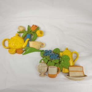 May include: Two decorative wall plaques featuring a yellow teapot, flowers, and teacups. One plaque includes a vase of tulips, while the other has a cake and a mug. The plaques are in shades of yellow, orange, blue, and green.
