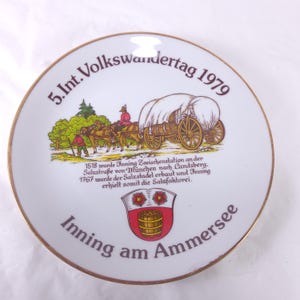 May include: A white decorative plate with a gold rim, featuring a colorful illustration of a horse-drawn wagon and text. The text includes "5. Int. Volkswandertag 1979" and "Inning am Ammersee".