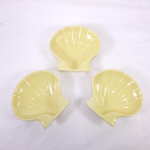 May include: Three pale yellow seashell-shaped dishes. The dishes have a glossy finish and are arranged on a white surface. The dishes are likely for serving snacks or small portions.
