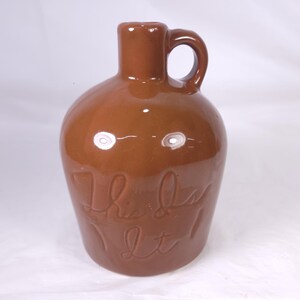 May include: A brown ceramic jug with a handle and the words "This is it" inscribed on the front. The jug has a rounded body and a narrow neck, with a glossy finish.