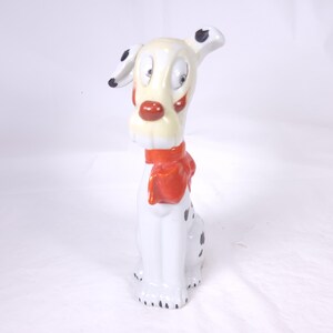 May include: A ceramic Dalmatian figurine with a cream-colored head, red nose, and a red scarf. The dog has black spots and black accents on its ears. The figurine is sitting upright and has a whimsical, cartoonish design.