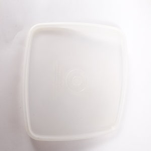 May include: A clear plastic square container lid with a raised circle in the center.