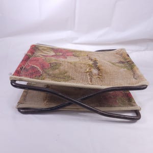 May include: A vintage folding footstool with a floral tapestry design in red, green, and beige. The tapestry is stretched over a dark metal frame. The footstool is folded, showing the underside of the tapestry.