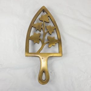 May include: A gold-colored trivet with a handle and a floral design. The trivet is shaped like an iron, with a handle at the bottom and a decorative cutout of a tree with leaves. The trivet is made of metal and is likely used to protect surfaces from hot items.