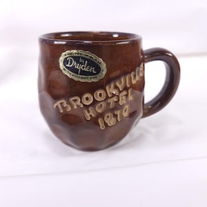 May include: A brown ceramic mug with a handle. The mug has the words "Brookville Hotel 1870" in raised gold lettering. A gold and black sticker on the mug reads "A Melody in Glaze by Dryden with Volcanic Ash."
