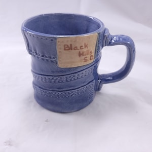May include: A blue ceramic mug with a denim-like design, featuring stitched details and a handle. A tan rectangular patch displays the text "Black Hills S.D."