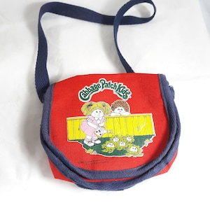 May include: Red canvas shoulder bag with a blue strap. The bag features a Cabbage Patch Kids logo and a graphic of two dolls watering a patch of cabbage.
