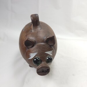 May include: A brown ceramic piggy bank with white painted details and green glass eyes. The piggy bank has a slot on the top for coins.