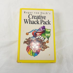 May include: A yellow box with the text "Roger von Oech's Creative Whack Pack" and a colorful illustration of a jester. The jester has a rainbow-colored mane and a hat with bells. The box is on a white surface.