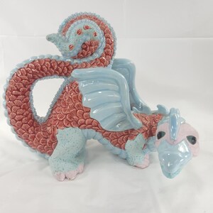 May include: A ceramic dragon figurine in shades of red, blue, and pink. The dragon has a long, curved tail, wings, and a detailed scale pattern. The figurine is a decorative item.